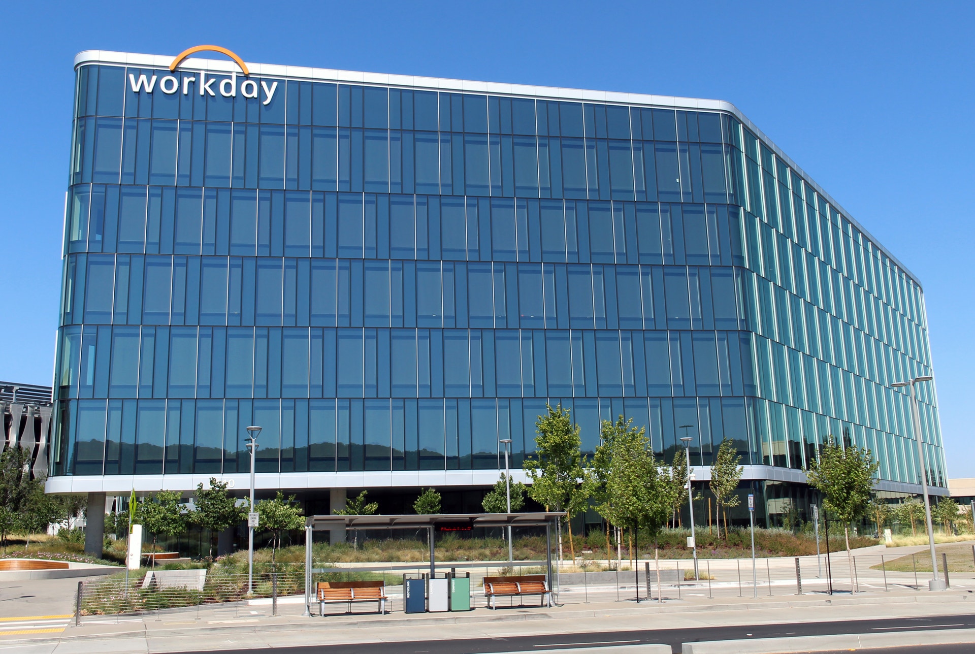 Workdays huvudkontor, en modern glasbyggnad med företagets logotyp på fasaden, omgiven av en öppen plats med träd och bänkar.