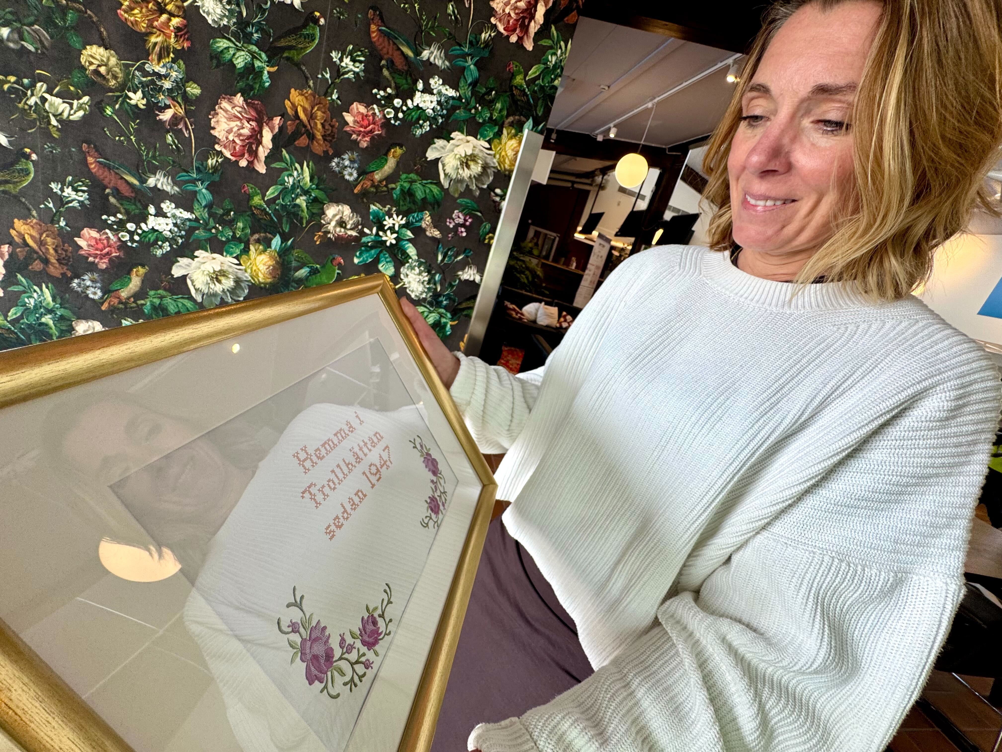 En kvinna håller upp en inramad blomsterillustrering med handskriven text.