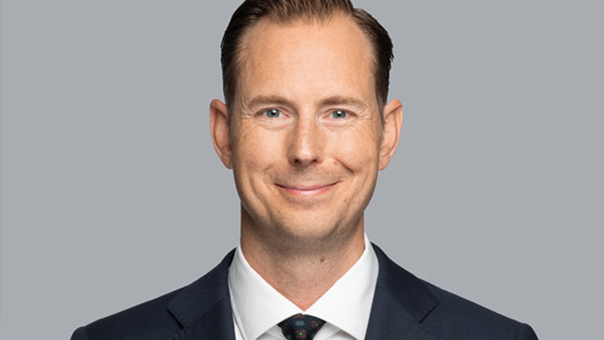 Porträtt av en man i mörk kostym och vit skjorta mot neutral grå bakgrund