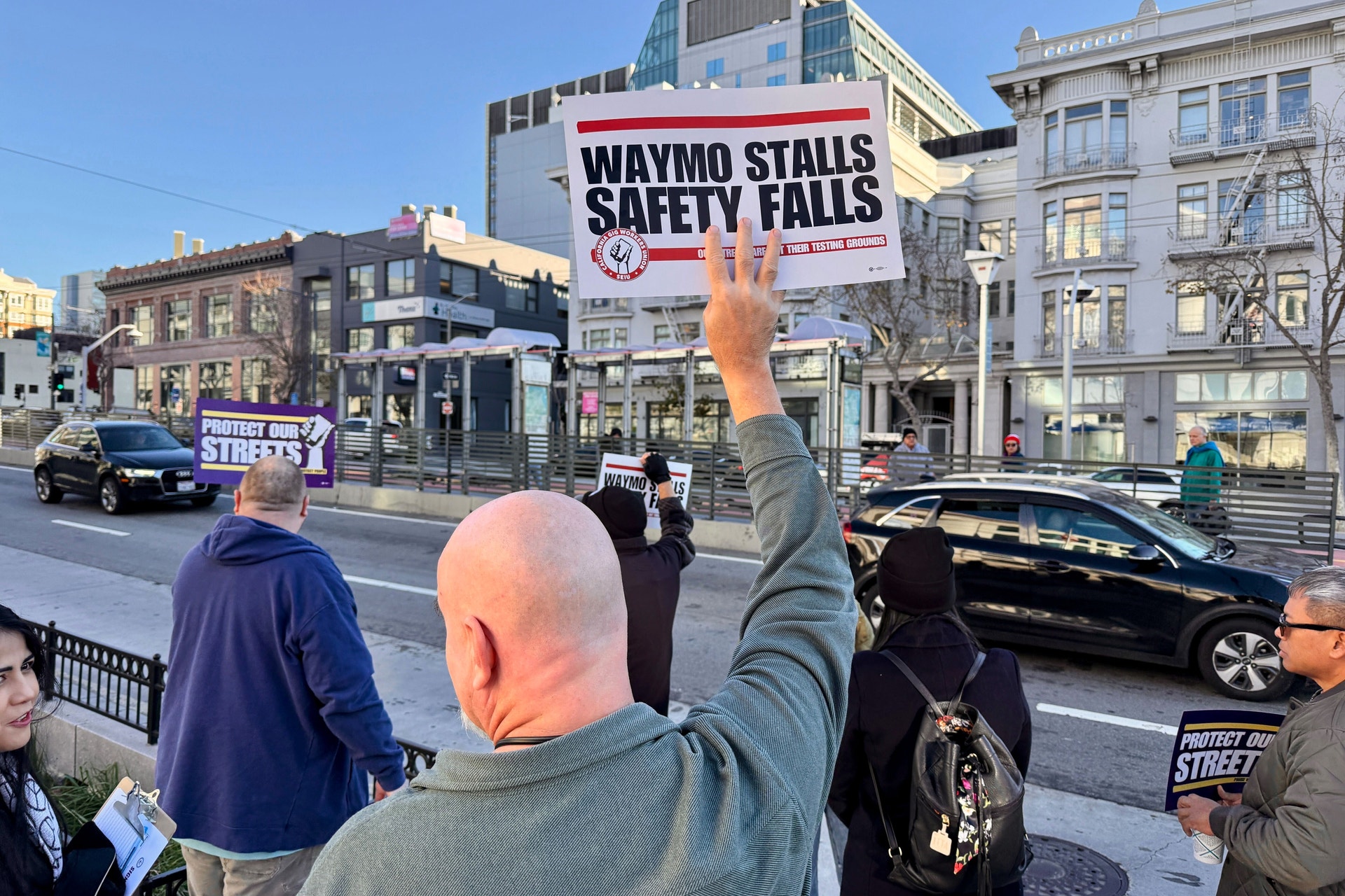 Gatudemonstranter håller upp protestplakat mot Waymo på en stadsgata på en solig dag.