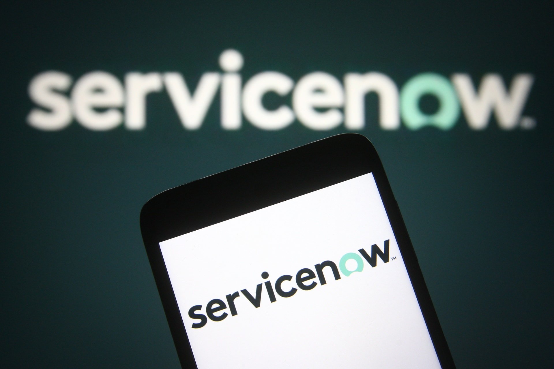ServiceNow-logotypen visas på en smartphone-skärm med logotypen även synlig i bakgrunden på en mörkblå yta.
