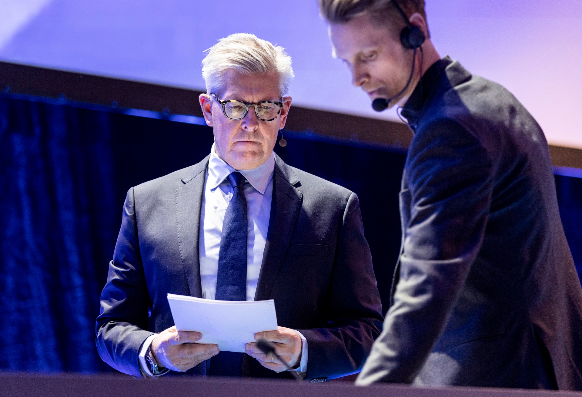 Två män på en scen under en presentation. En äldre man i kostym och glasögon håller i ett papper, och en yngre man med headset står bredvid honom.