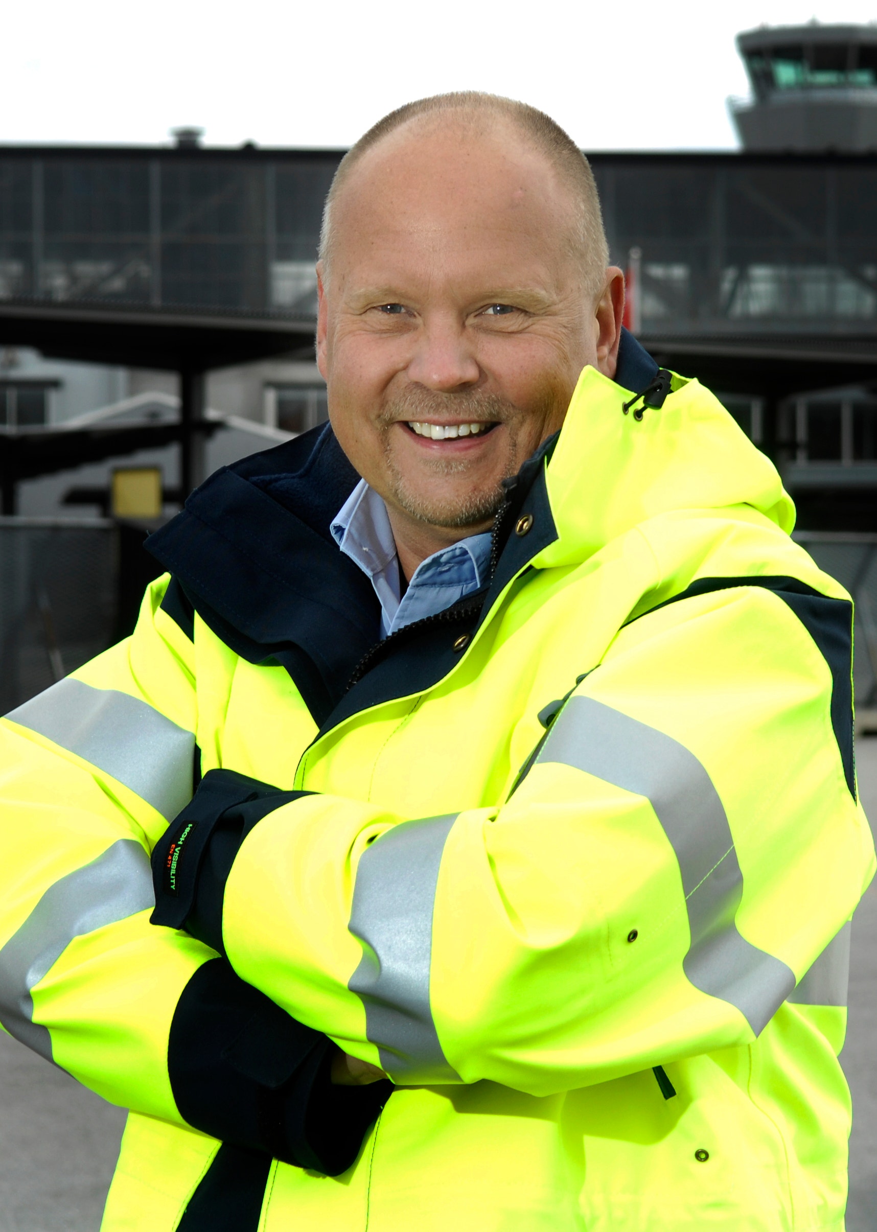 En leende man i gul varseljacka står med armarna i kors utomhus vid en flygplats.