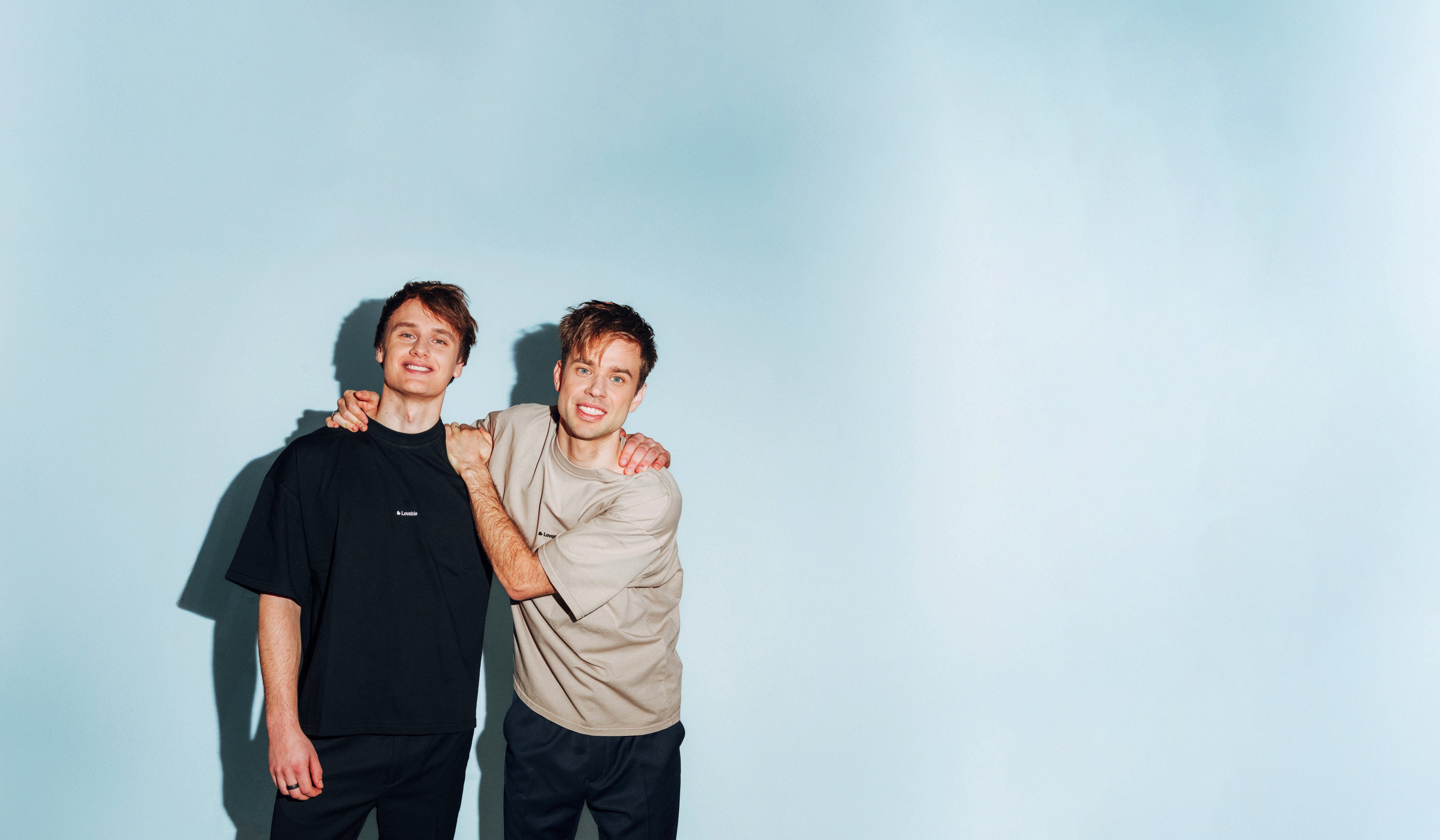 Två unga män står tillsammans och ler mot kameran framför en ljusblå bakgrund.