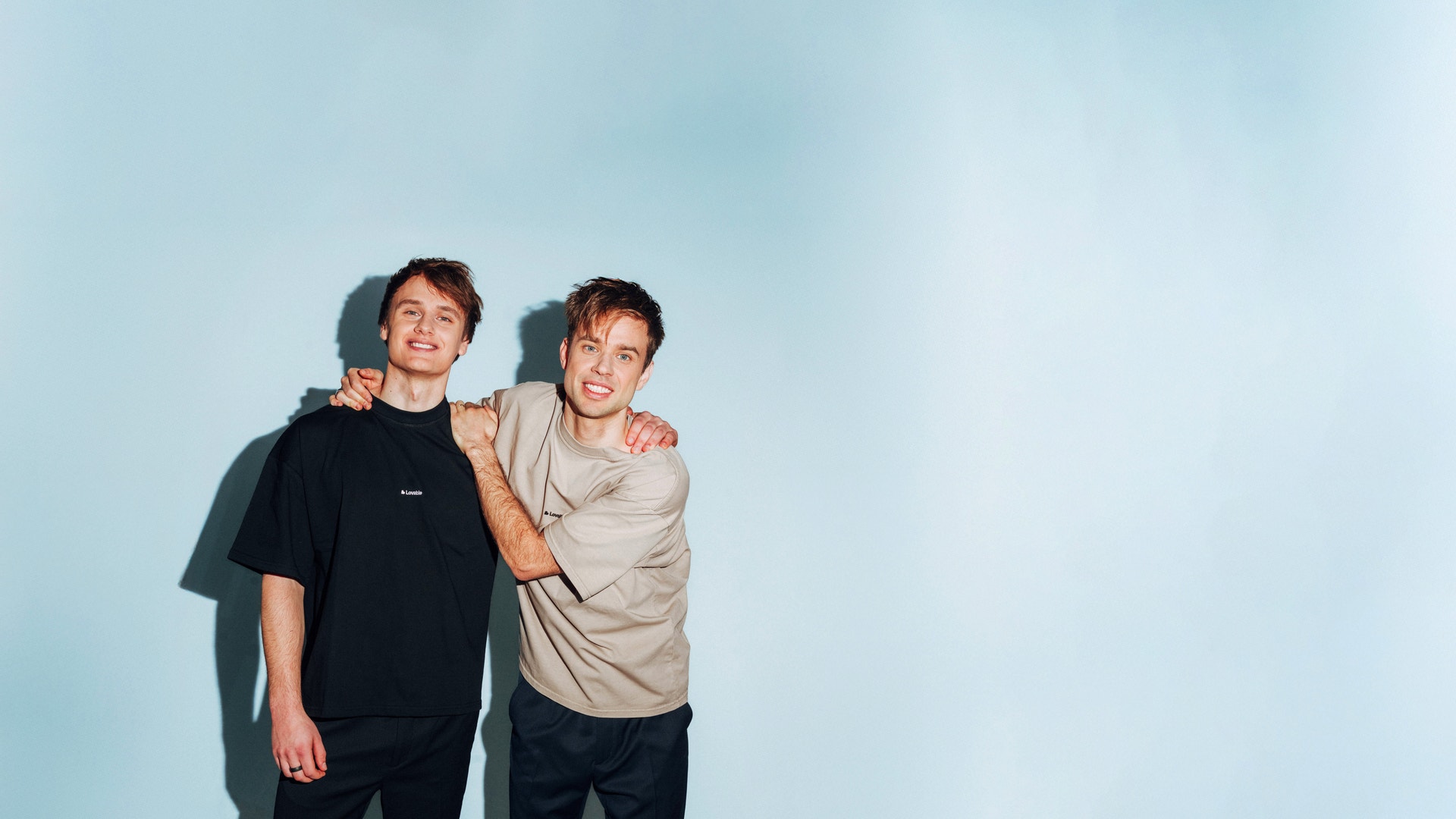 Två unga män står tillsammans och ler mot kameran framför en ljusblå bakgrund.