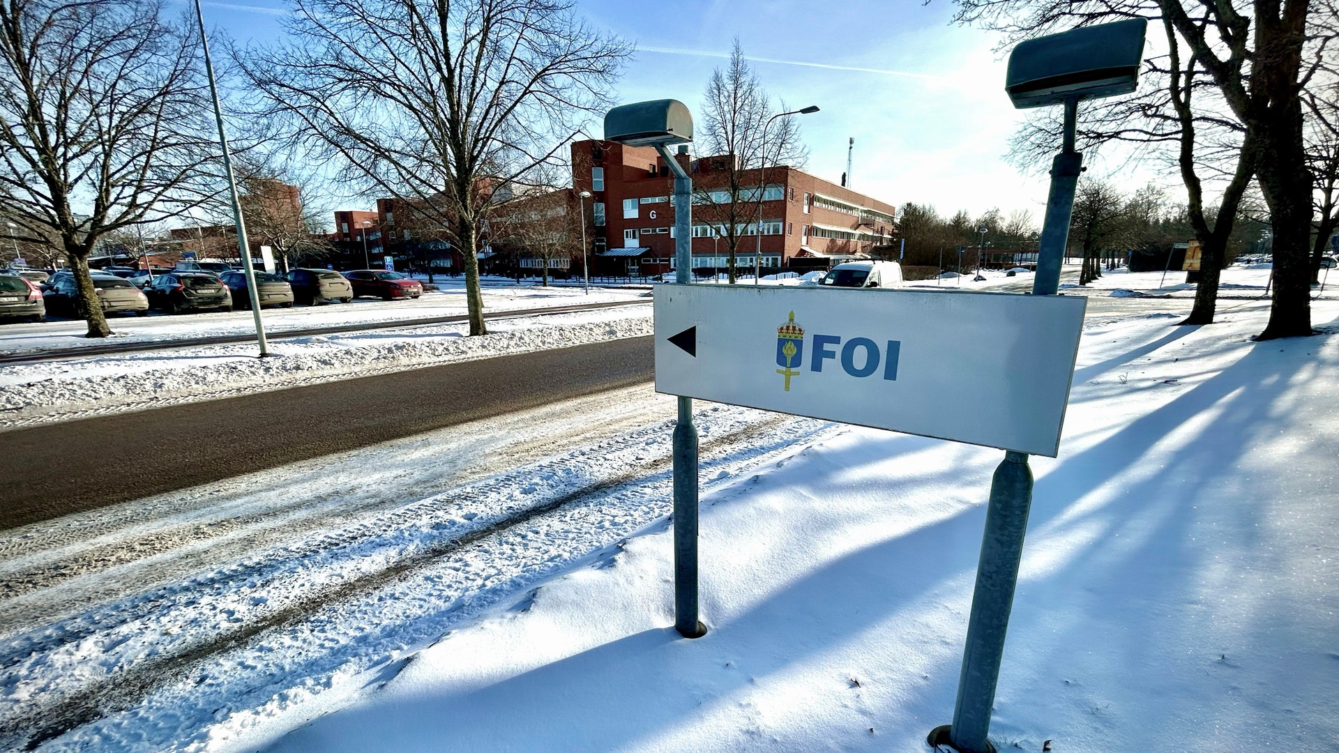 En vit vägvisare med FOI-logotypen i ett snötäckt parkområde med träd och byggnader i bakgrunden.