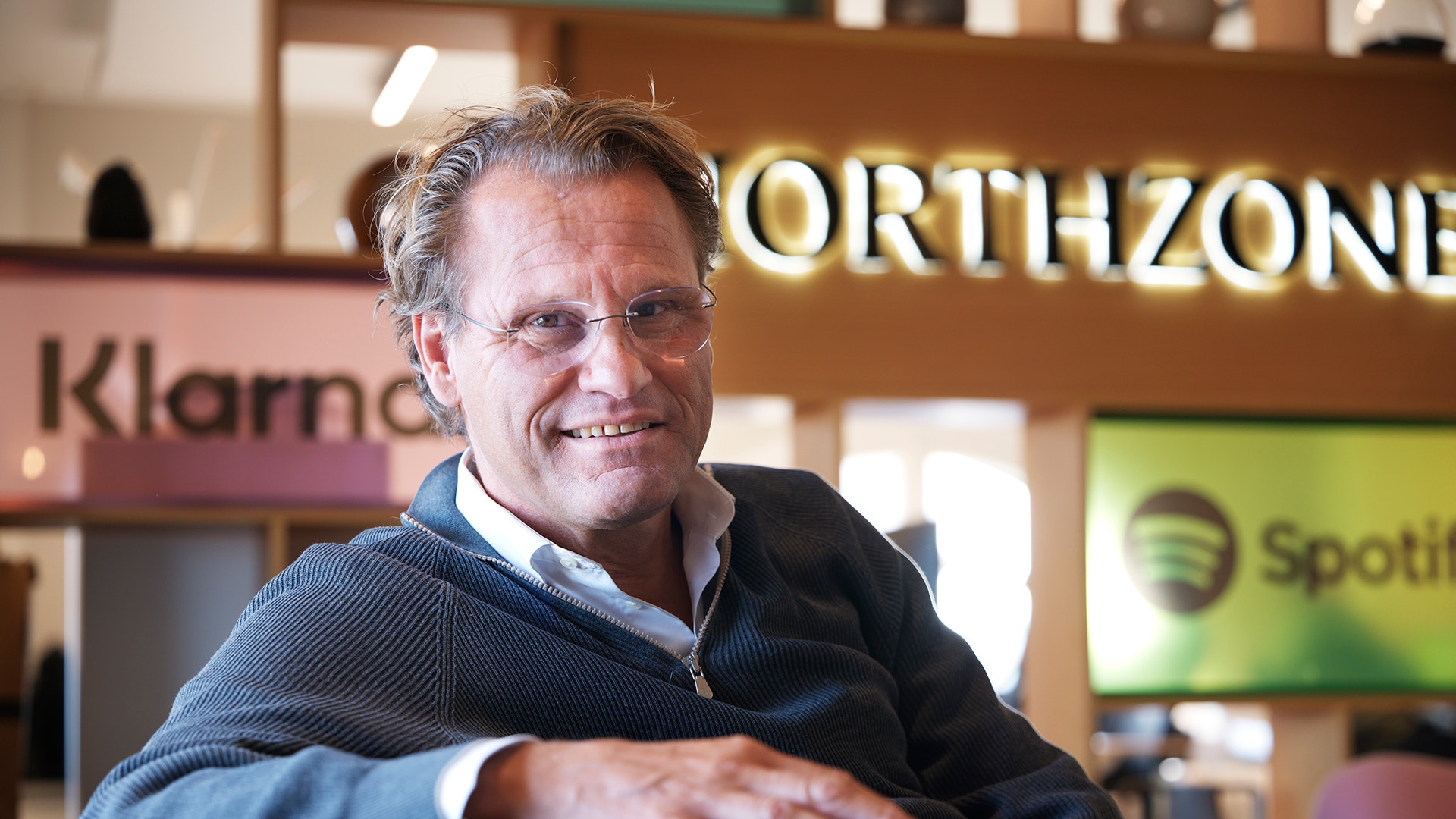 En man i stickad tröja ler mot kameran och lutar sig mot en grön soffa, med Northzones kontor i bakgrunden.