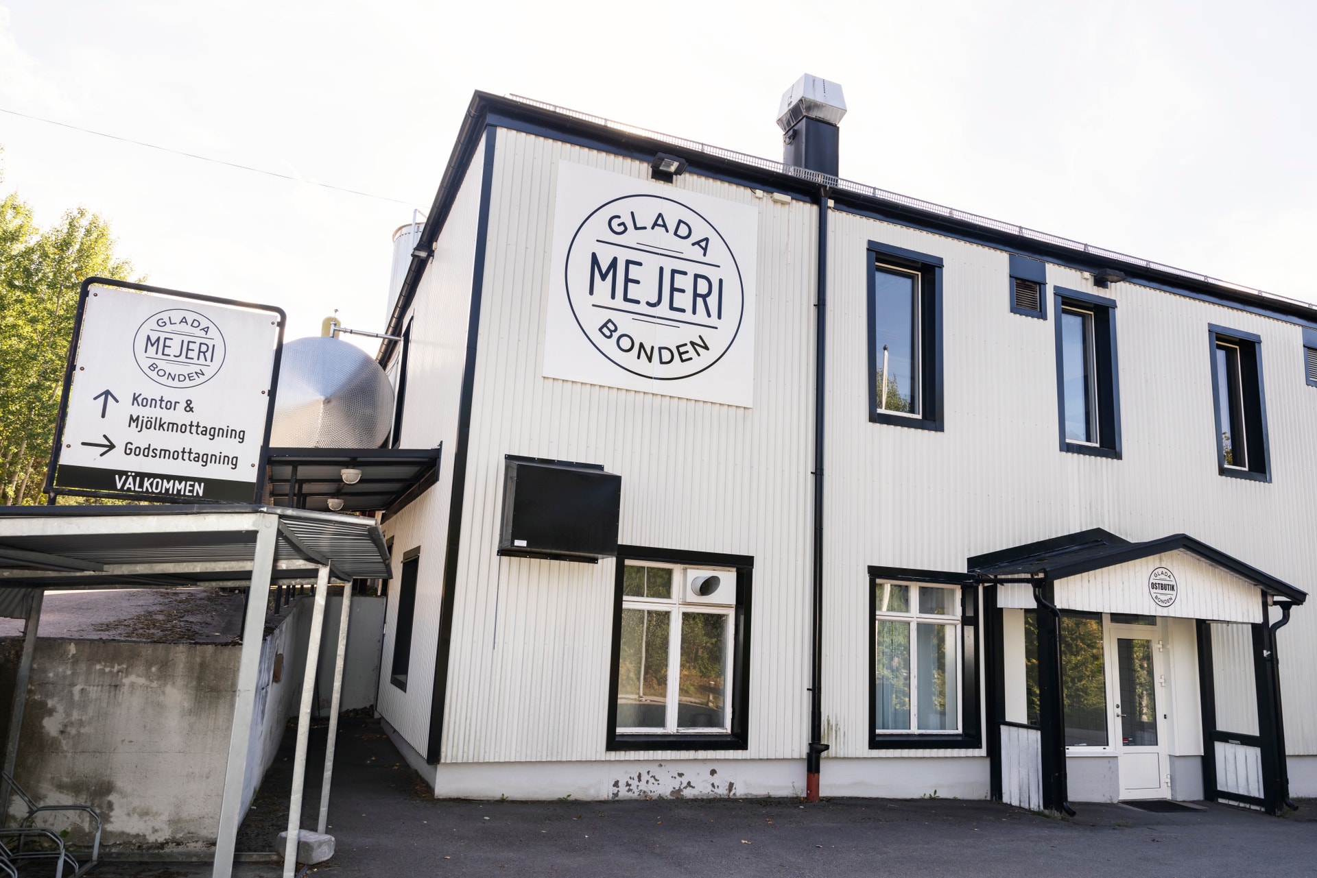 Vit byggnad med svarta fönster och detaljer. Logotyp för Glada Bonden Mejeri på fasaden. Skylt vid entrén till höger.
