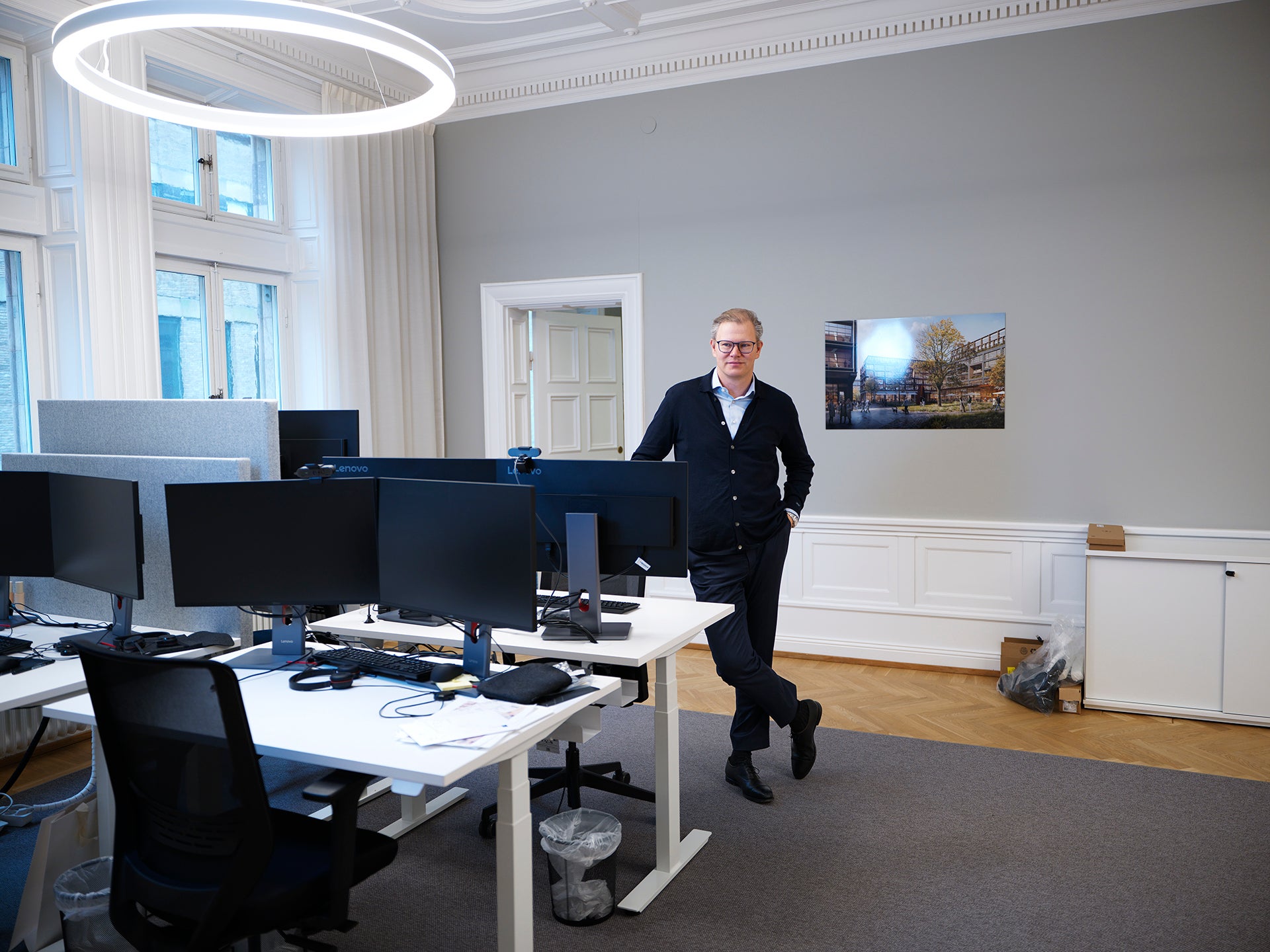 En man i mörk kofta står lutad mot ett skrivbord i ett ljust kontor med flera arbetsplatser.