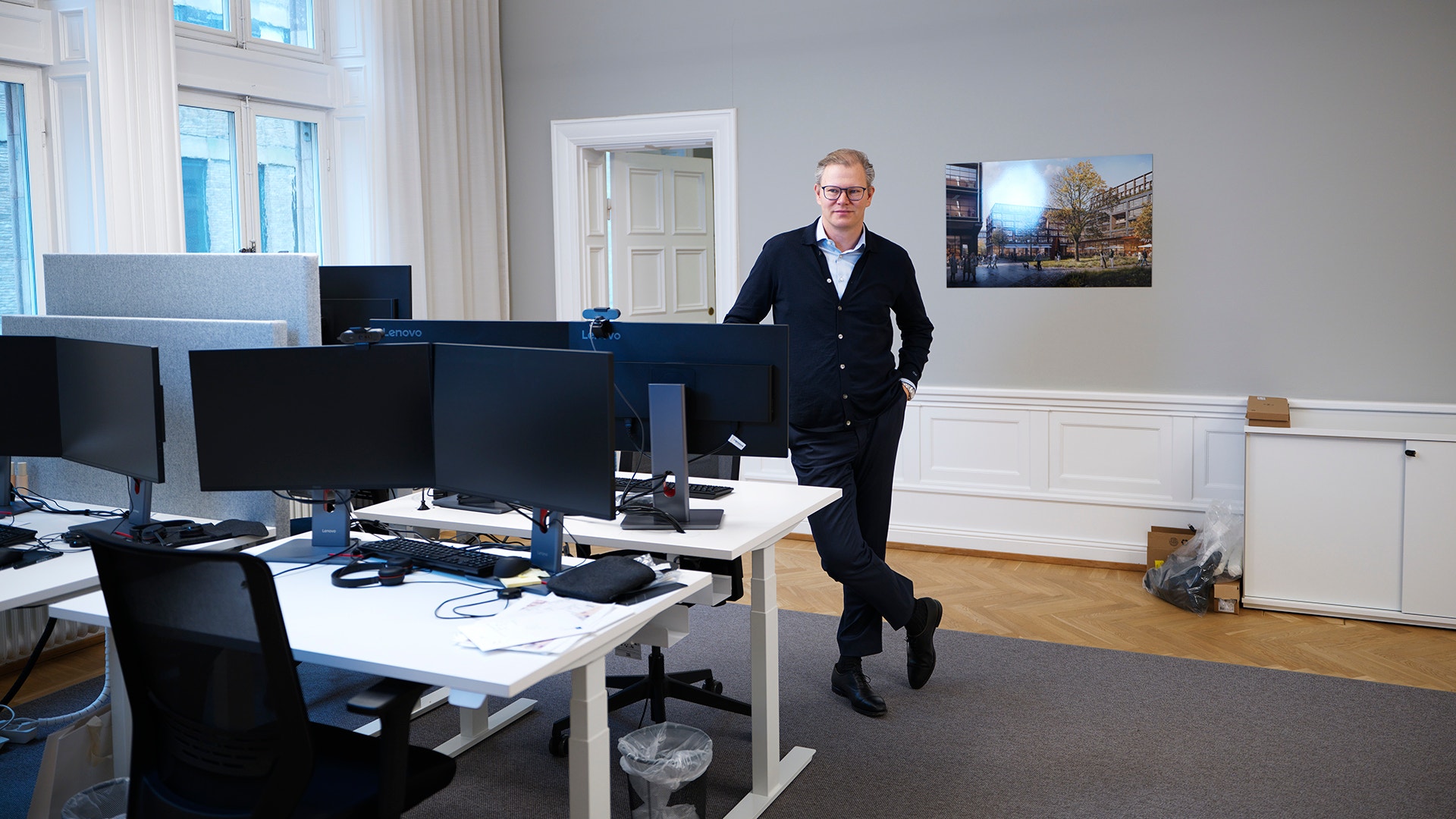 En man i mörk kofta står lutad mot ett skrivbord i ett ljust kontor med flera arbetsplatser.