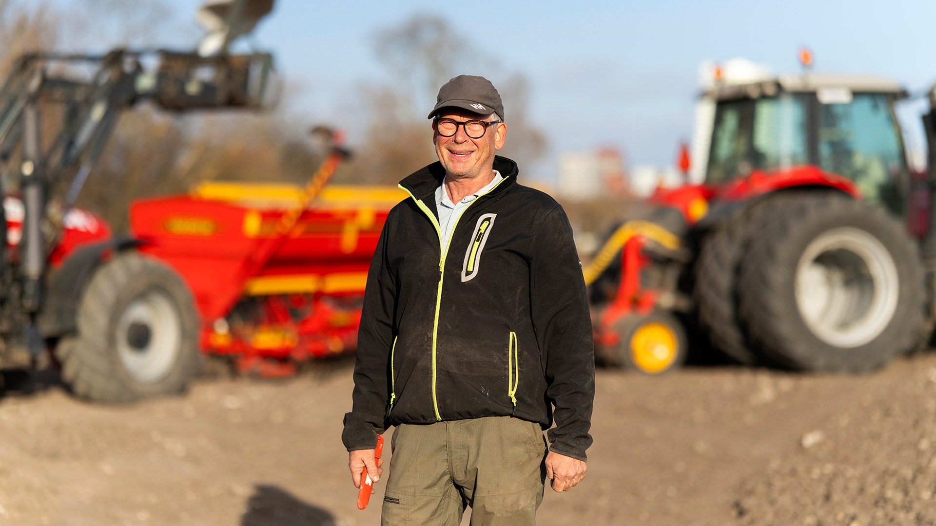 Man i svart jacka och keps står framför lantbruksmaskiner och traktorer på en åker.