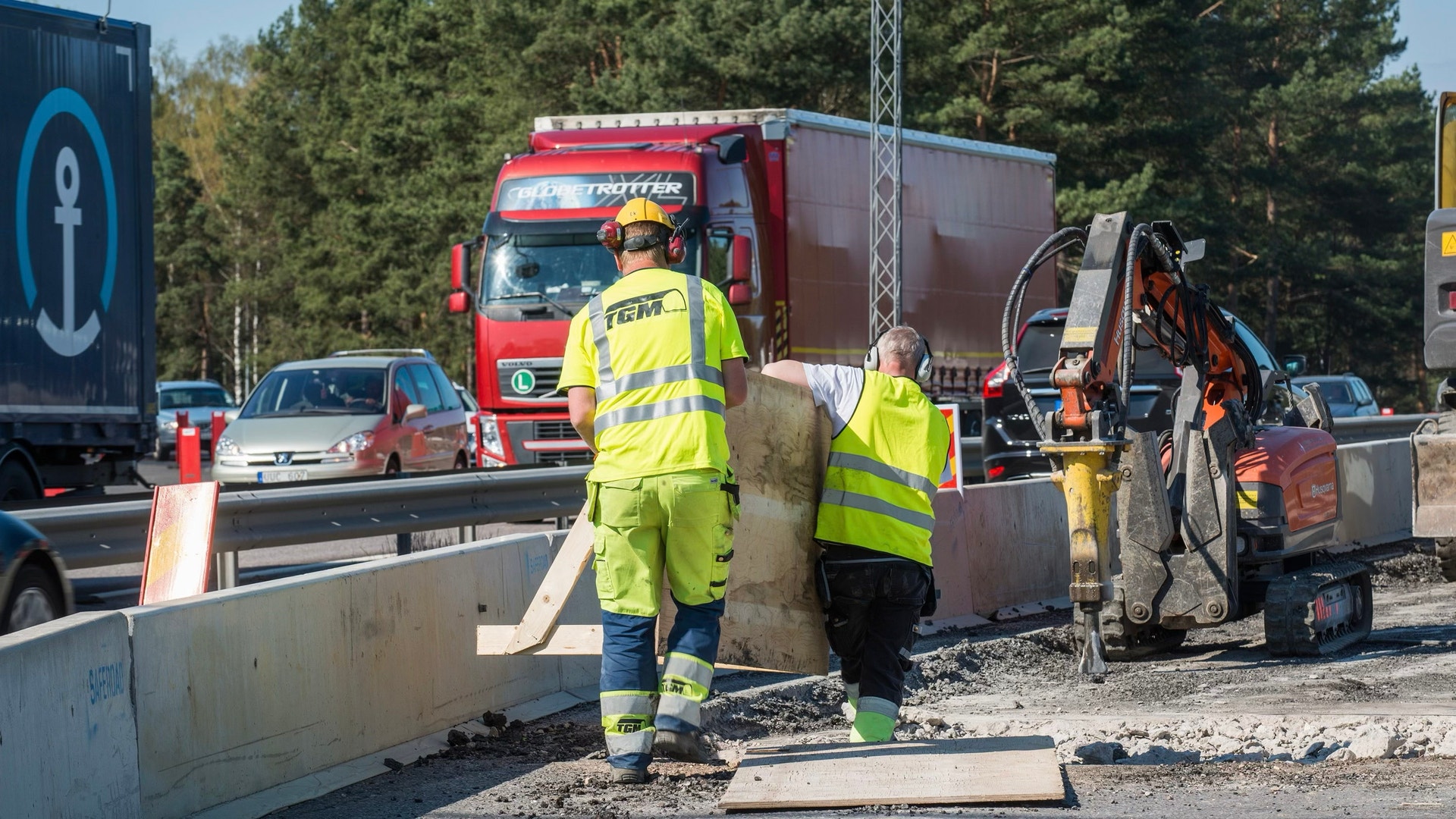 Arbetare i gula västar arbetar vid vägkonstruktion på E4-vägen. En röd lastbil och olika byggmaskiner syns i bakgrunden.