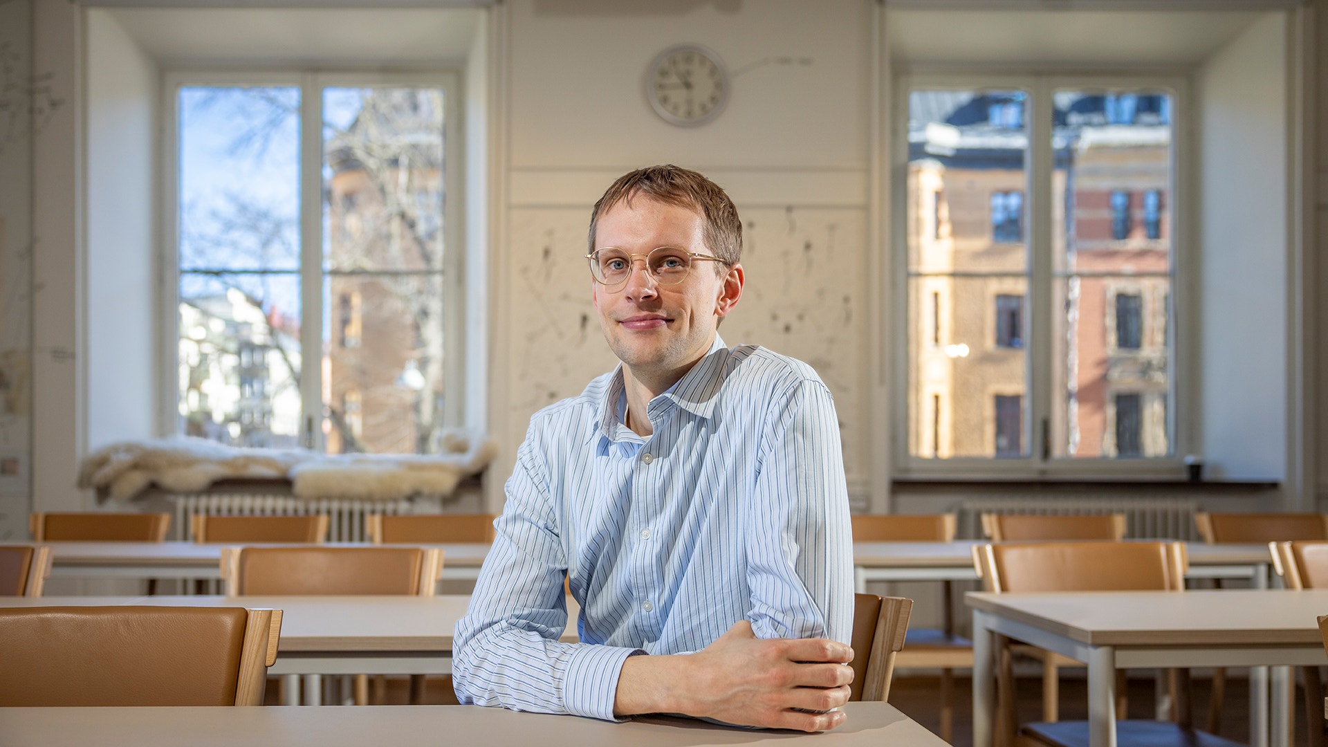 En man i glasögon och ljusblå randig skjorta sitter vid ett bord i ett klassrum med stora fönster.