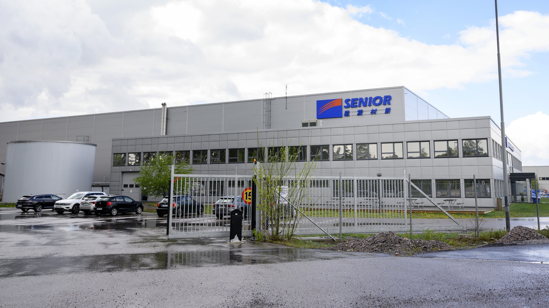 En grå industribyggnad med Senior-logotyp på fasaden. Parkeringsplats med bilar framför byggnaden och metallstaket vid entrén.