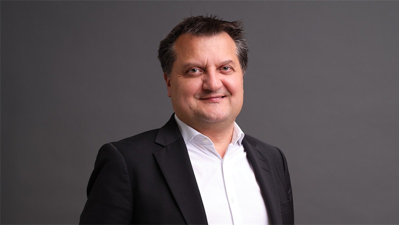 Porträtt av en man i mörk kostym och vit skjorta mot grå bakgrund