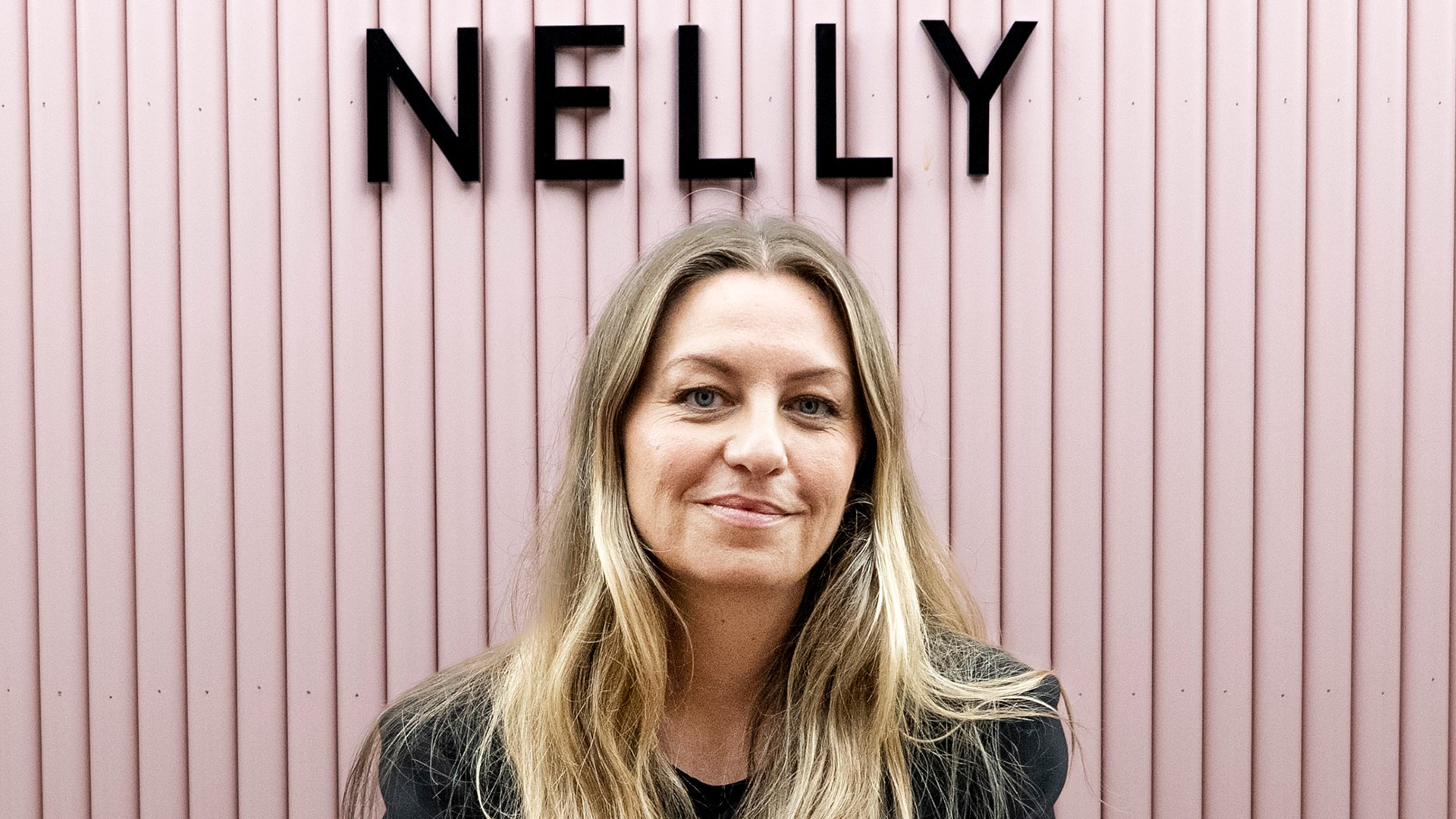Porträttfoto av en kvinna med långt blont hår som ler mot kameran framför en rosa vägg med texten "NELLY"