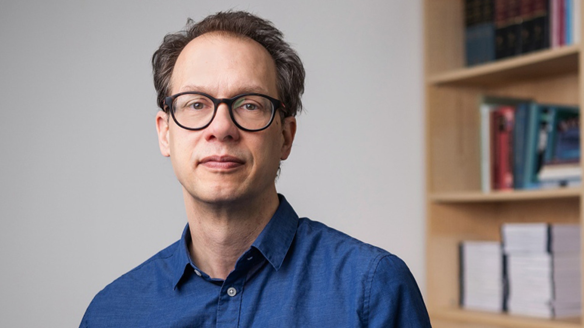 Porträtt av Jonas Vlachos, man med glasögon i blå skjorta framför bokhylla