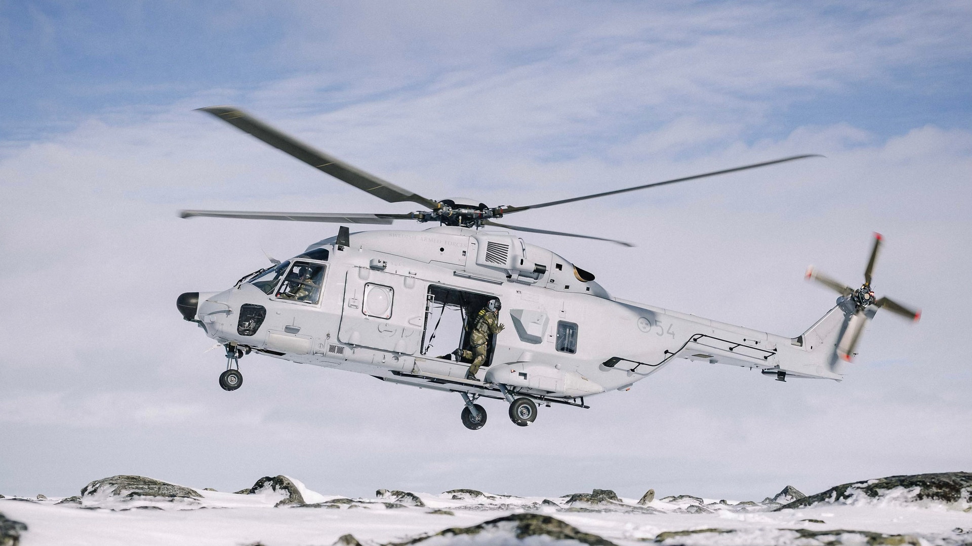 En vit militär helikopter flyger över ett snötäckt fjälllandskap under molnig himmel.