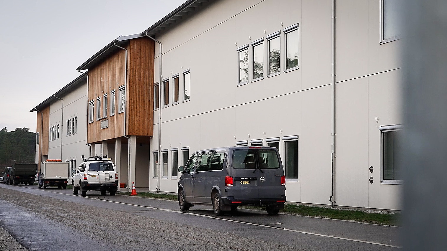 Modern byggnad med vita och grå väggar, trädetaljer och stora fönster. Två bilar är parkerade på vägen framför byggnaden.