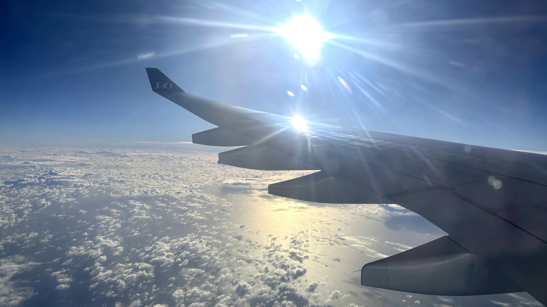 Flygplansvinge tillhörande flygbolaget SAS, sett från fönstret under flygning, med sol och molnlandskap i bakgrunden.