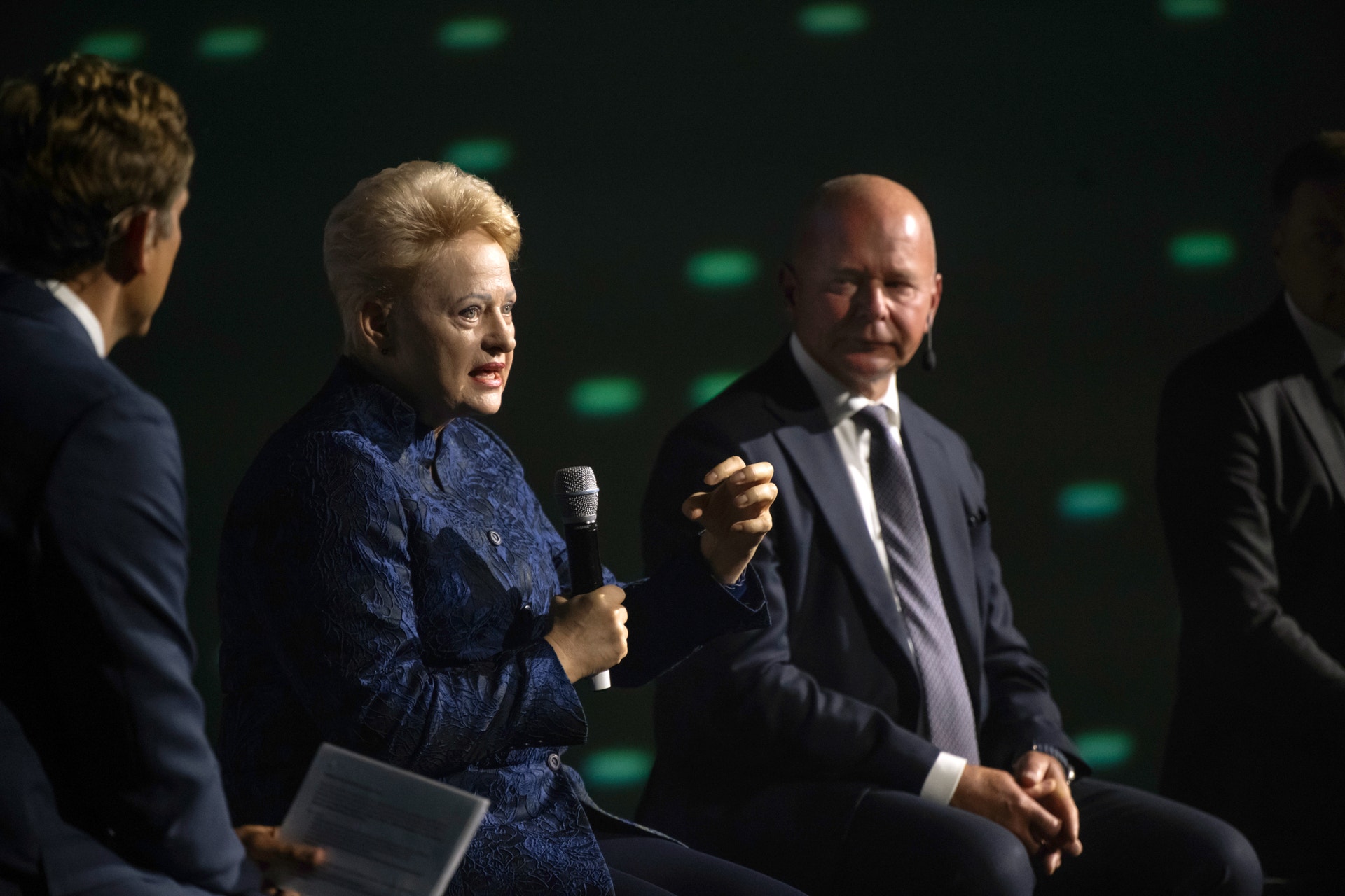 En äldre kvinna i mörkblå klänning håller i en mikrofon och talar under en paneldiskussion på en scen med tre män i kostym. Grön bakgrund med ljuseffekter.