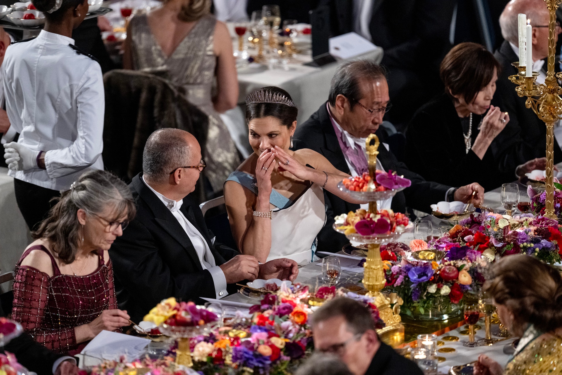 Gäster sitter vid ett dekorerat festbord med färgglada blommor och äter under en formell gala eller festlig sammankomst.