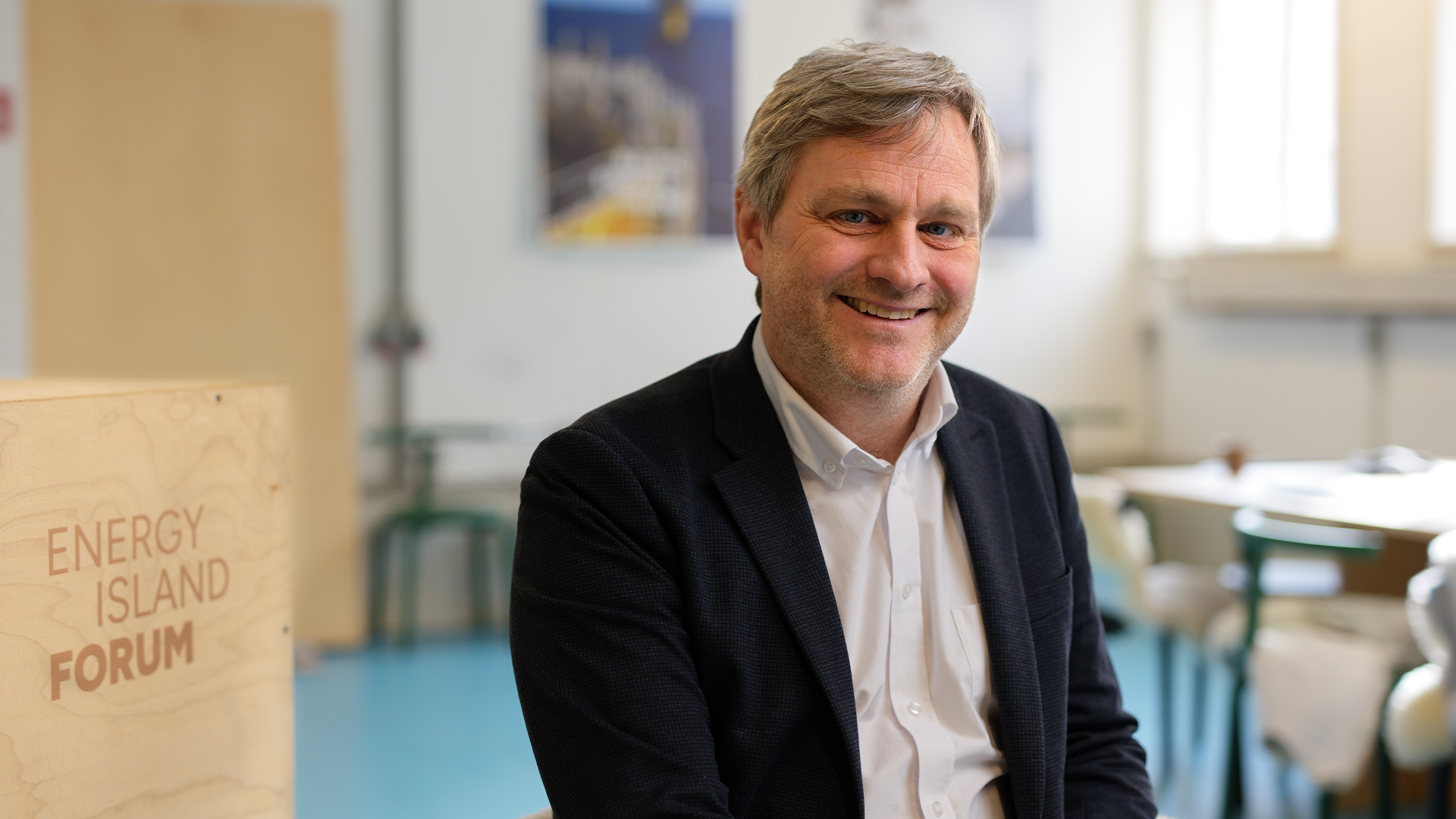 Porträtt av en man i mörk blazer och vit skjorta som ler mot kameran i ett kontorsmiljö.