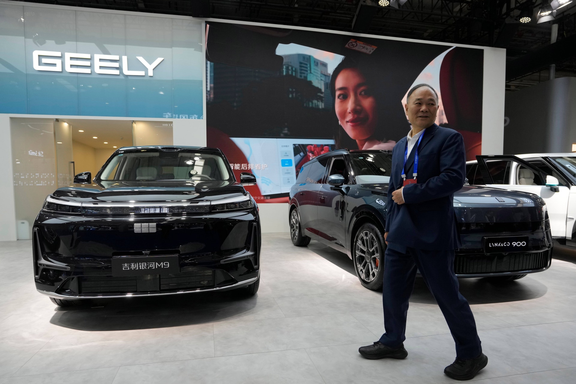 En man i mörk kostym står vid en bilmässa framför flera svarta Geely-bilar med Geely-logotypen synlig i bakgrunden.