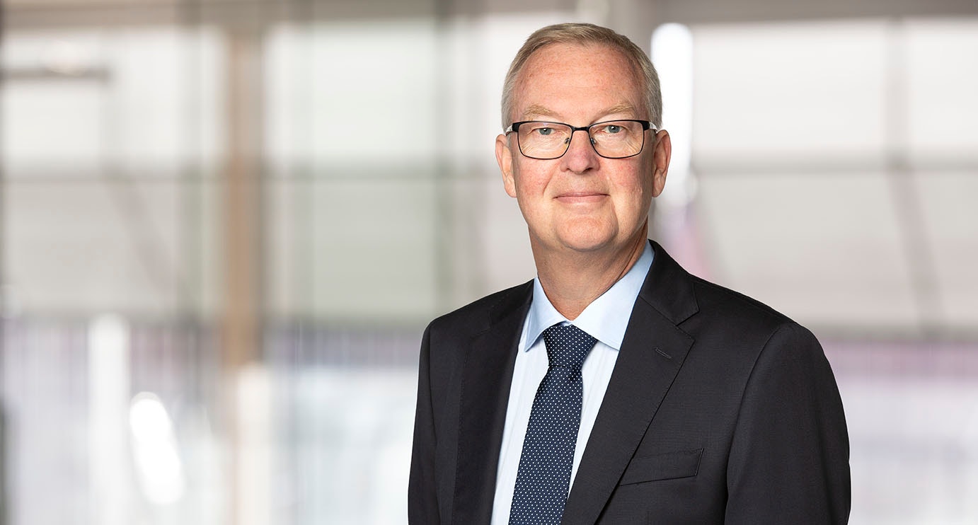 Porträtt av en man i svart kostym och blå slips mot en suddig kontorsbakgrund.