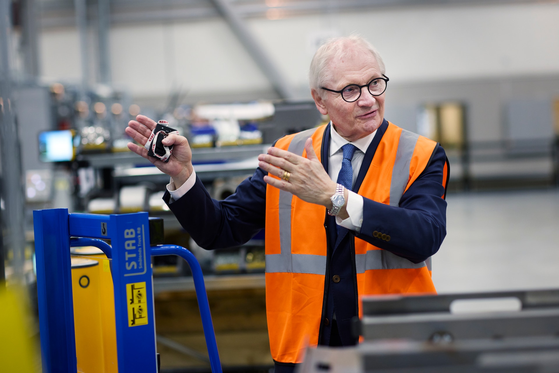 En äldre man i säkerhetväst och kostym står i en fabrik och presenterar en maskinkomponent.