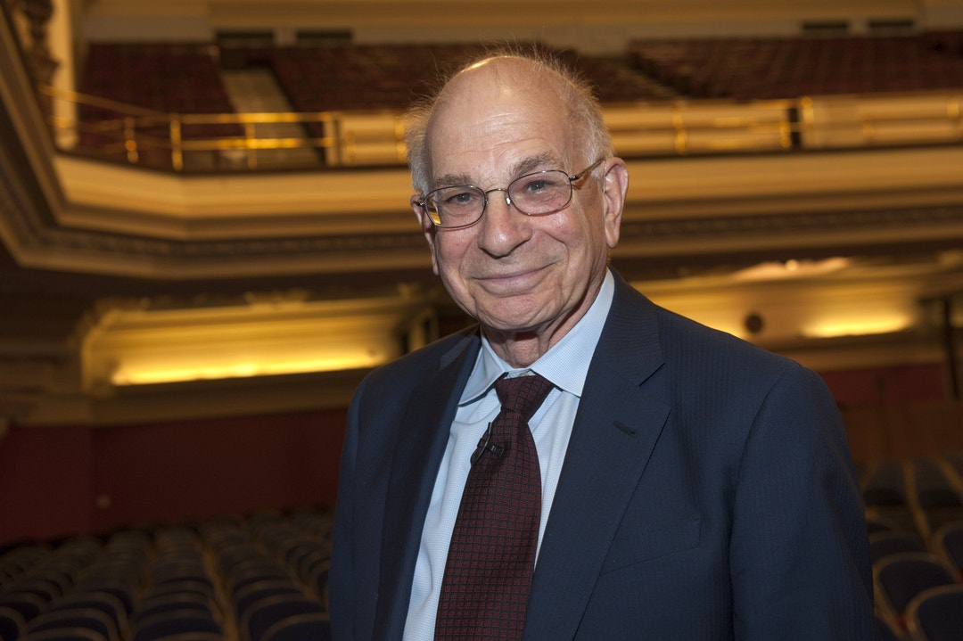 En äldre man i kostym och slips ler mot kameran i en teater- eller konsertsalong.