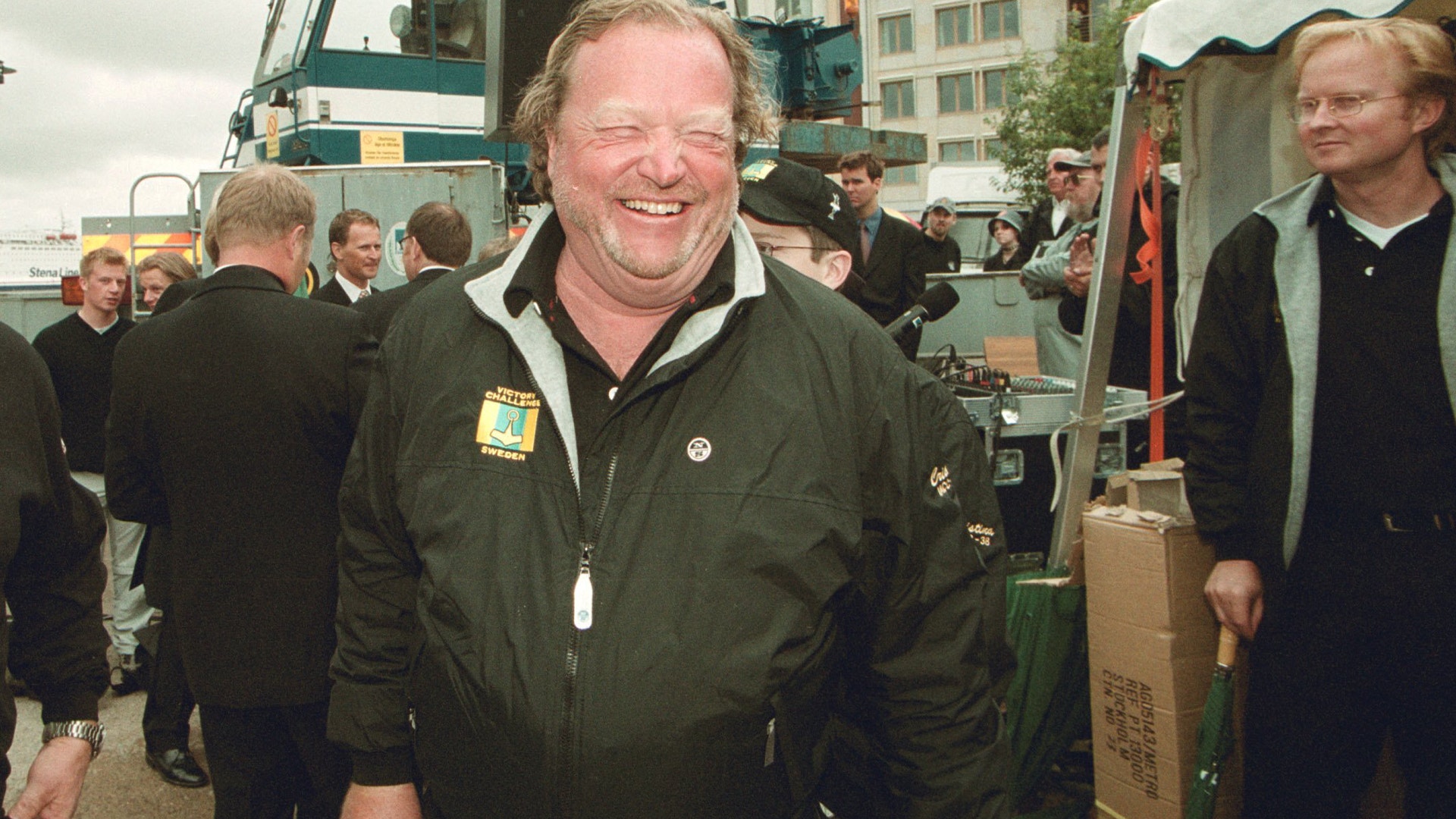 En man i grön jacka ler mot kameran, omgiven av andra människor på en utomhusplats.