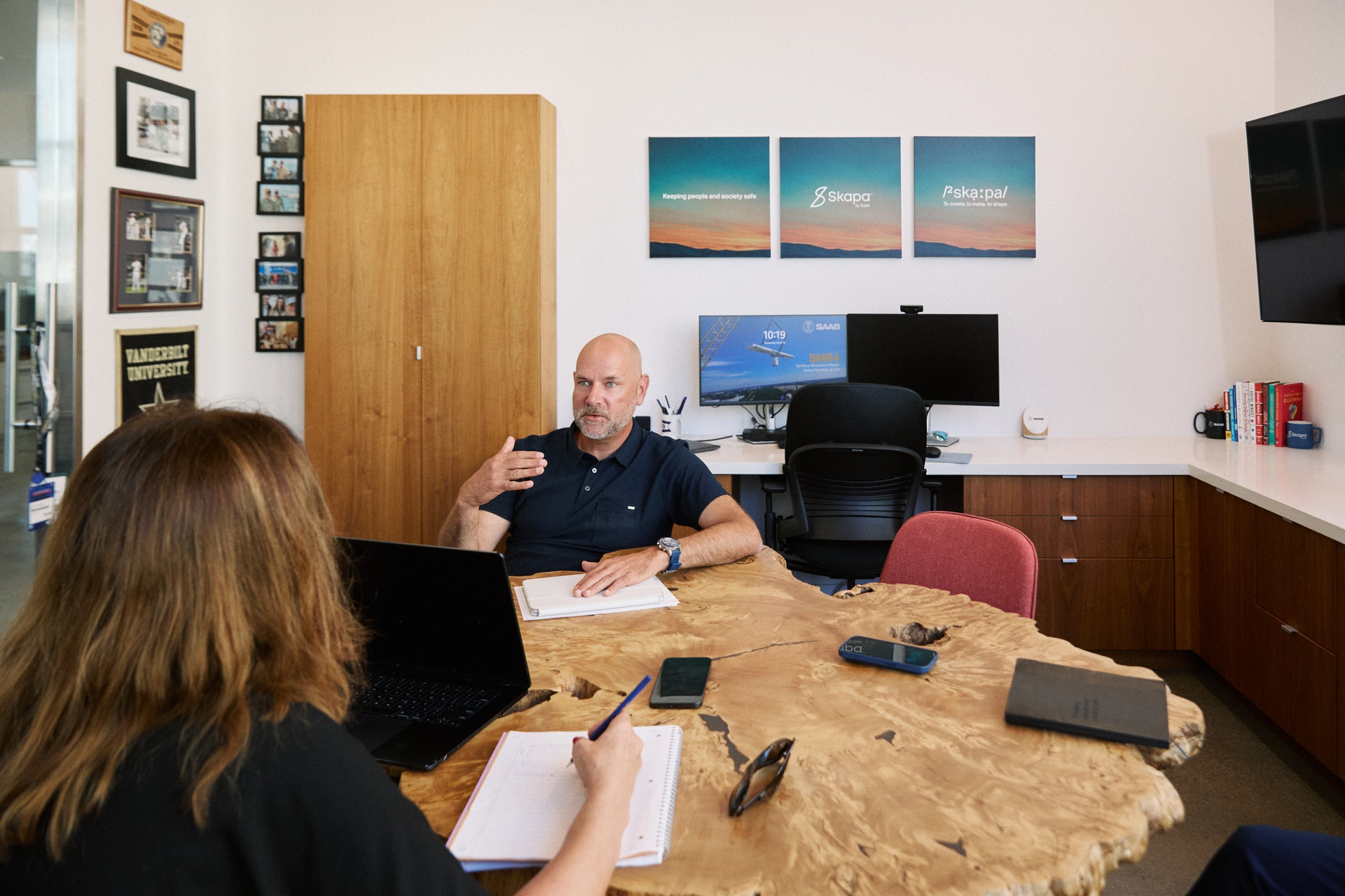 En man och en kvinna sitter vid ett träbord på ett kontor och diskuterar. Mannen gestikulerar medan han pratar, och kvinnan lyssnar med anteckningar framför sig.