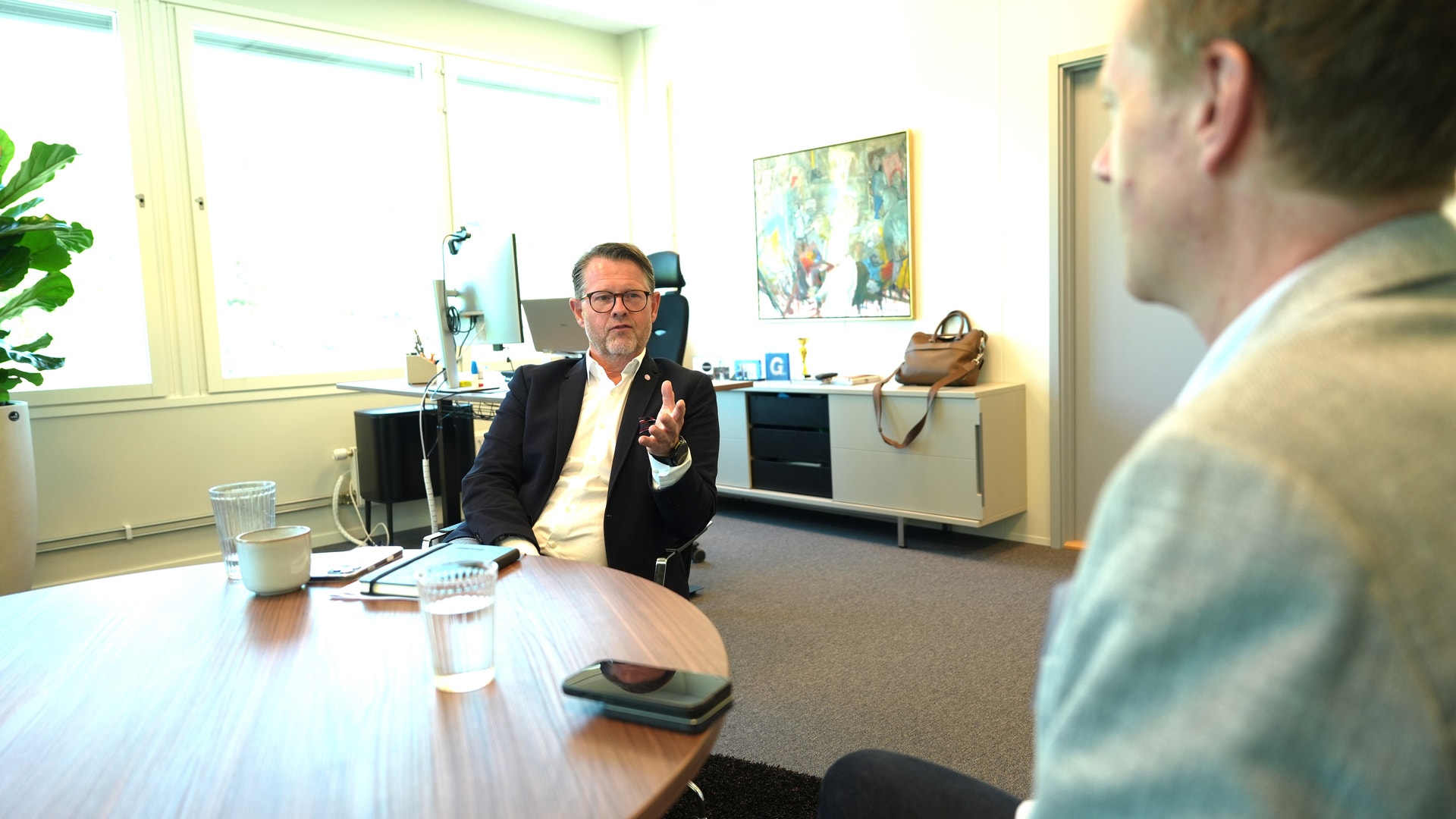 Två män i ett modernt kontor sitter vid ett bord och diskuterar. Mannen i mörk kostym gestikulerar medan han talar.