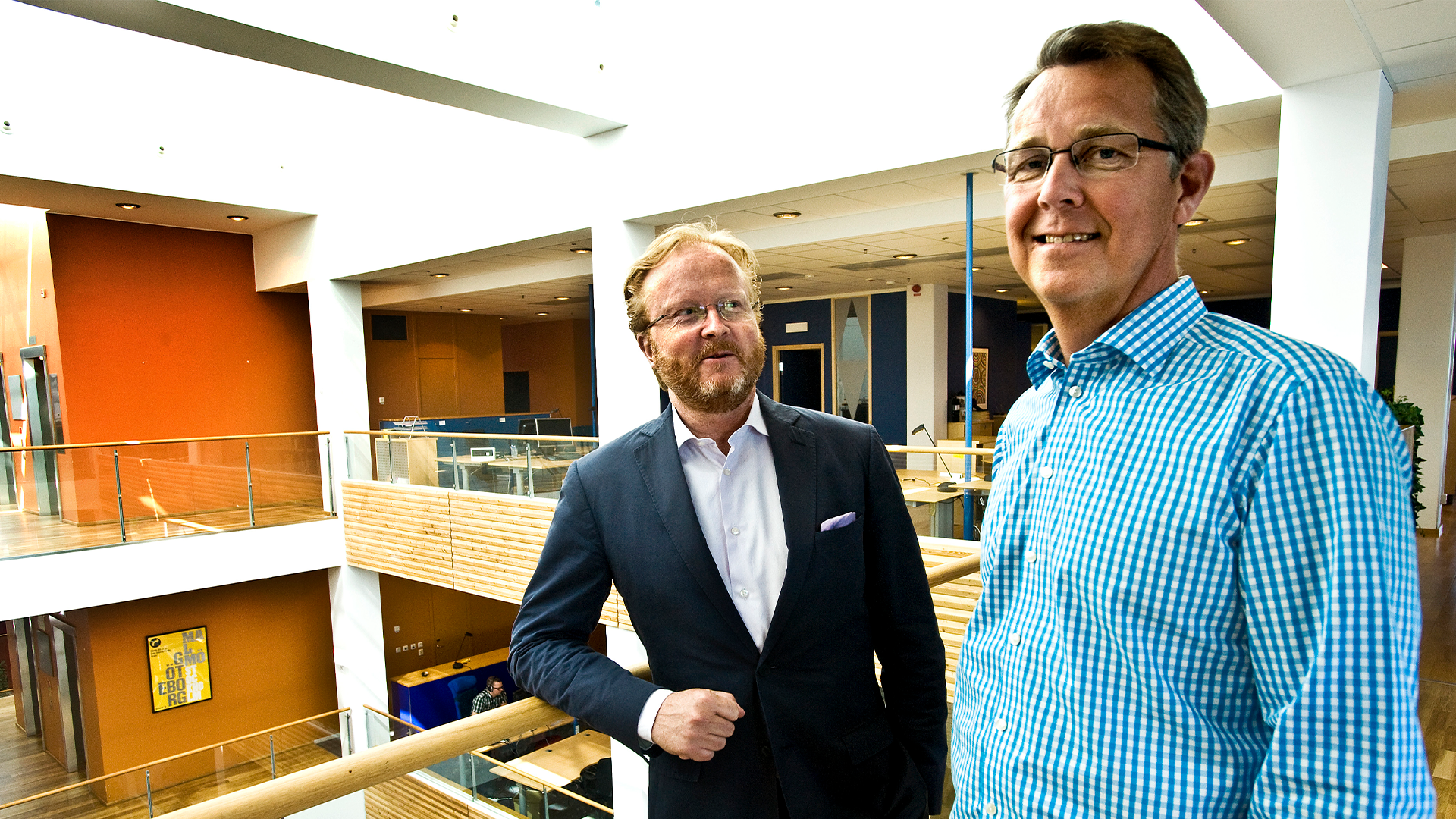 Två män står tillsammans i en modern byggnad. Mannen till vänster bär mörk kostym och vit skjorta, mannen till höger bär ljusblå rutig skjorta. I bakgrunden syns orange väggar och moderna arkitektoniska element.