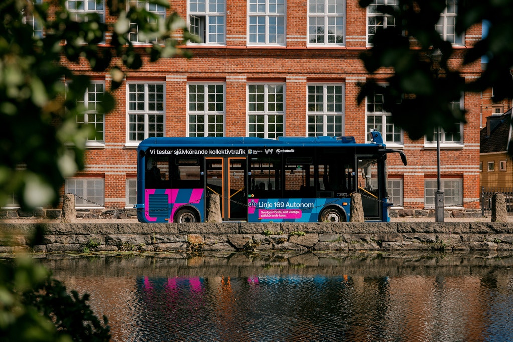 En svart buss med rosa och blå detaljer parkerad vid en kanal framför en röd tegelbyggnad.