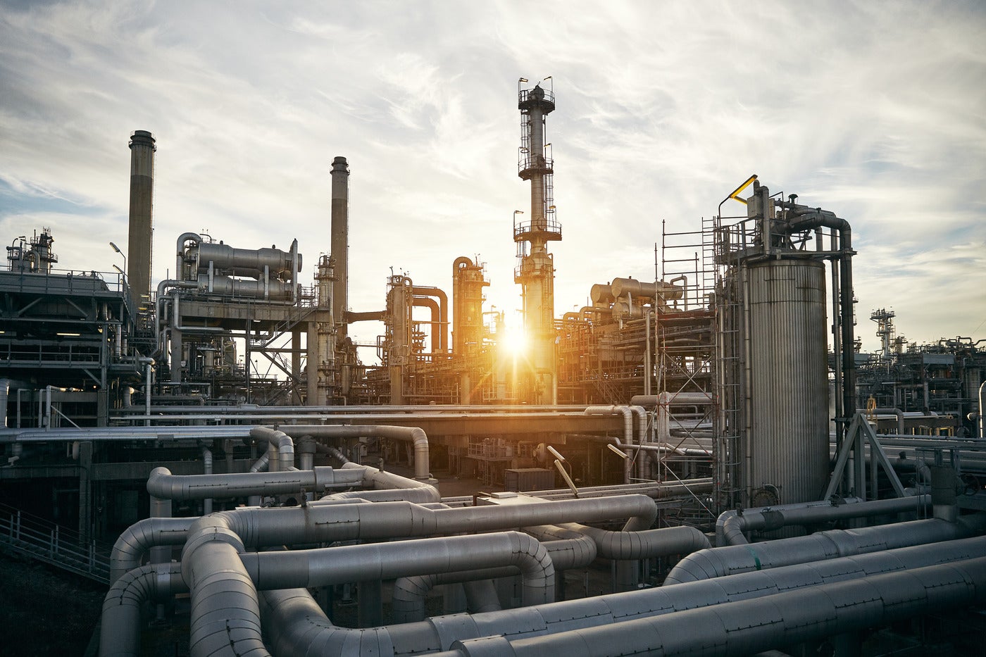 En stor raffineringsanläggning med industriella rör, torn och utrustning mot en molnig himmel vid solnedgång.