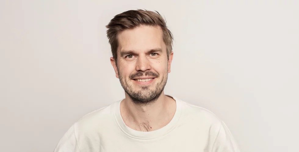 Porträtt av en man med skägg som ler mot kameran. Han bär en vit t-shirt och fotograferas mot en ljus bakgrund.