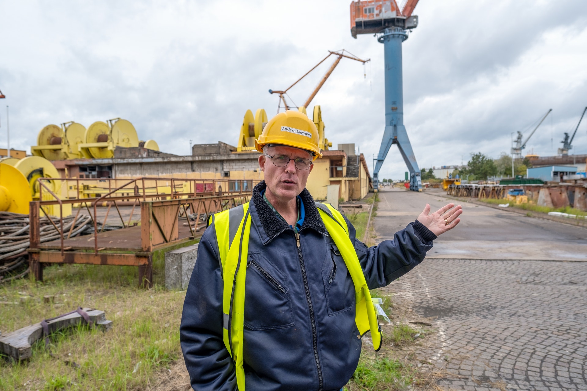 Man i gul säkerhetshjalm och reflektvest står i en industriell hamnanläggning med gul utrustning och kranar i bakgrunden.